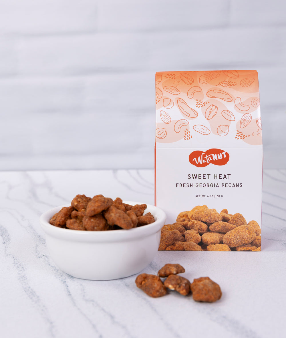 A box of WataNut Sweet Heat Fresh Georgia Pecans stands next to a small white bowl filled with sweet Heat flavored  pecans on a light marble surface against a white brick background.