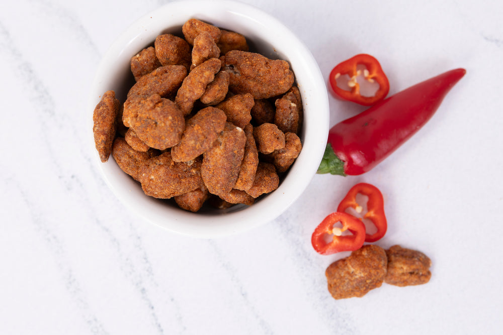 A white bowl filled with roasted Pecans coated in a sweet and spicy topping, styled on a white marble surface next to a whole red chili pepper and two sliced red chili peppers.
