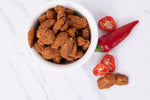 A white bowl filled with roasted Pecans coated in a sweet and spicy topping, styled on a white marble surface next to a whole red chili pepper and two sliced red chili peppers.