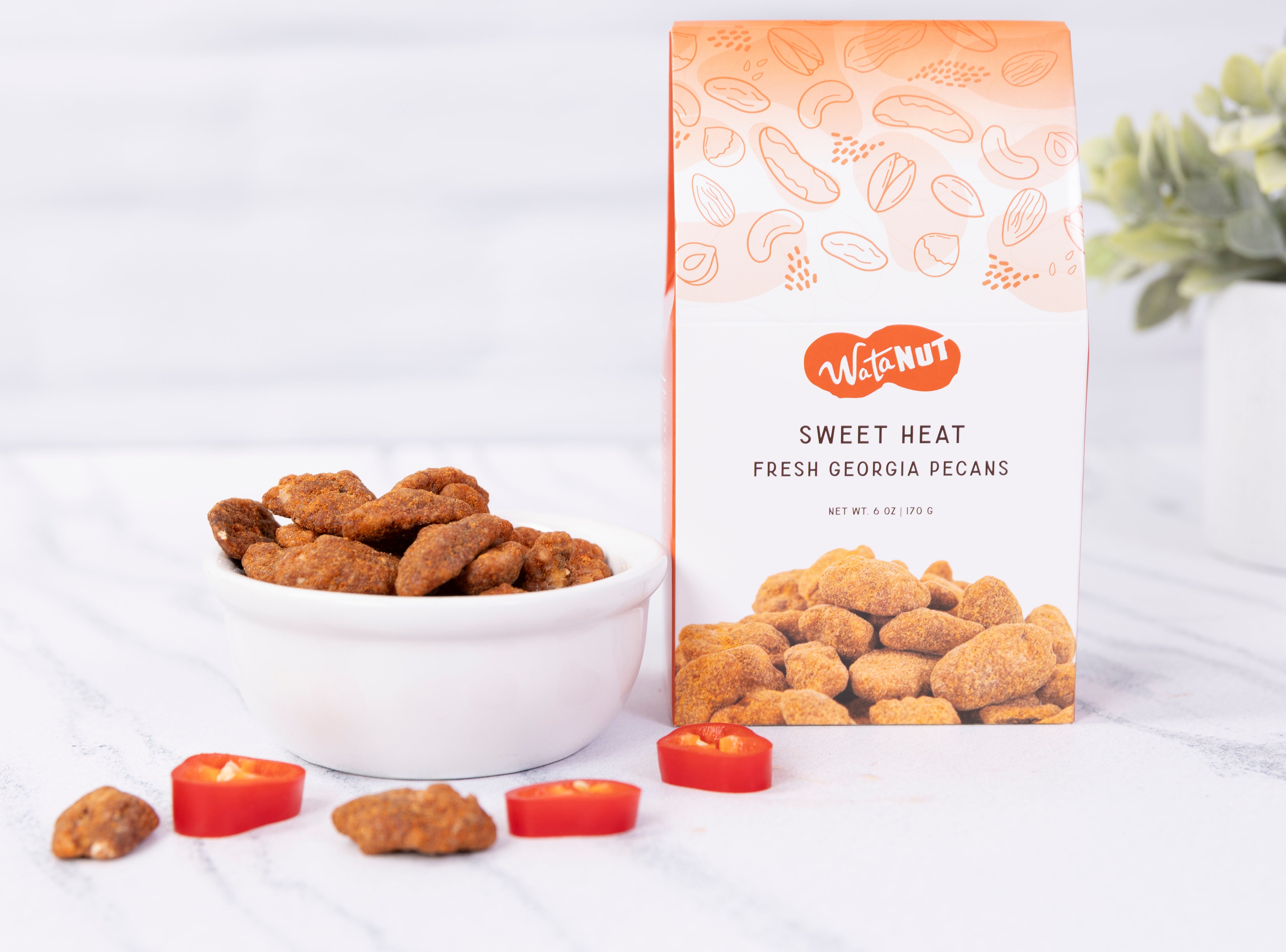 A bag of WataNut Sweet Heat Fresh Georgia Pecans stands next to a white bowl filled with the sweet and spicy pecans and a few sliced red chilies on a light marble surface.
