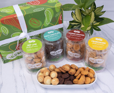 A WatanNut large cookie box featuring short bread cookies tubes in Key lime, Salty Chocolate, Cheddar Cocktail, and Cranstachio flavors, arranged on a white marble surface with a green patterned box behind it and a leafy plant to the right and a white dish of assorted cookies in front.
