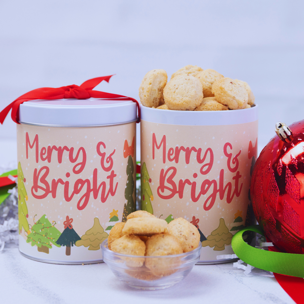 Two 'Merry & Bright' cookie tins with cookies and Christmas decorations on a light background