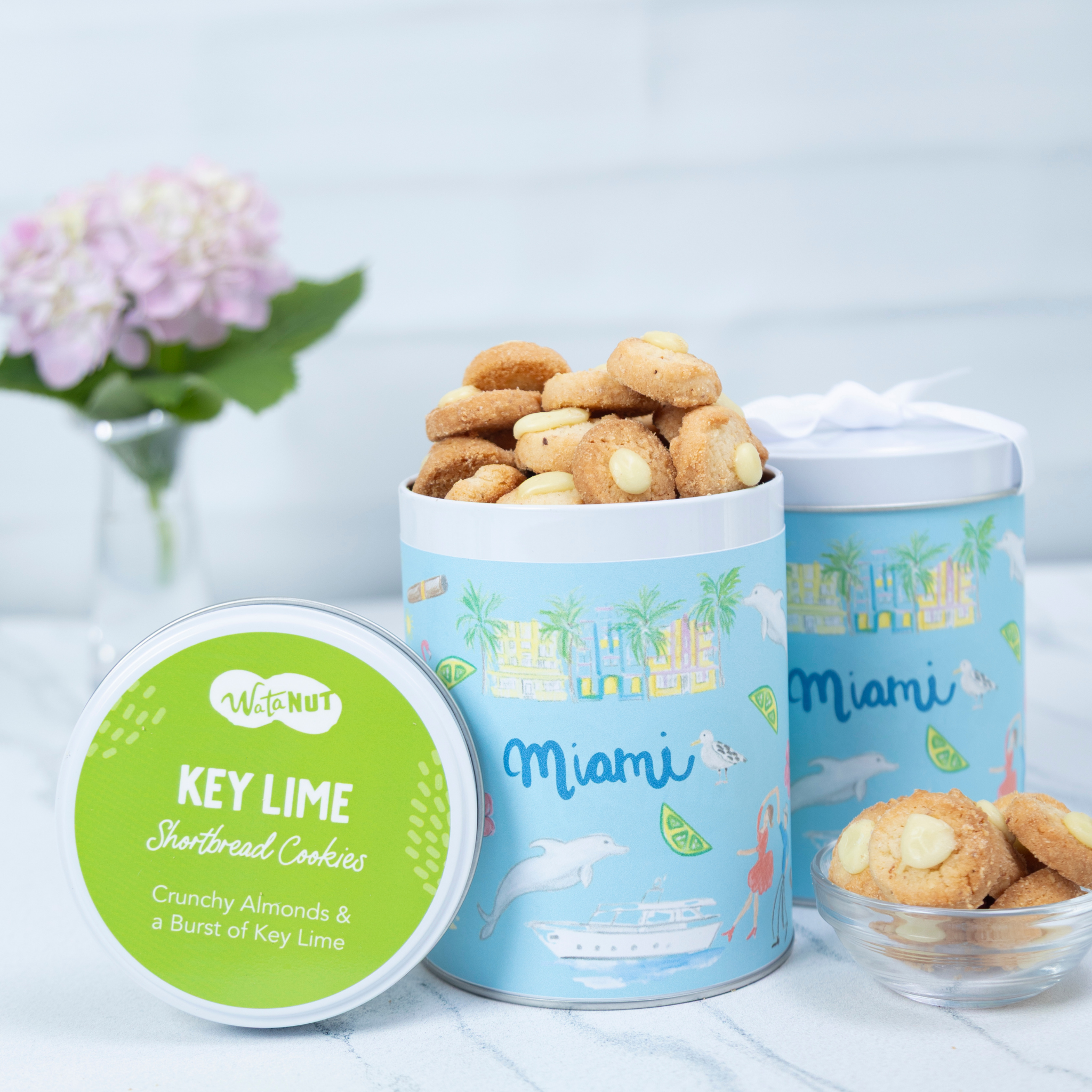 Key lime shortbread cookies in a blue tin with 'Miami' design, surrounded by flowers on a light background.